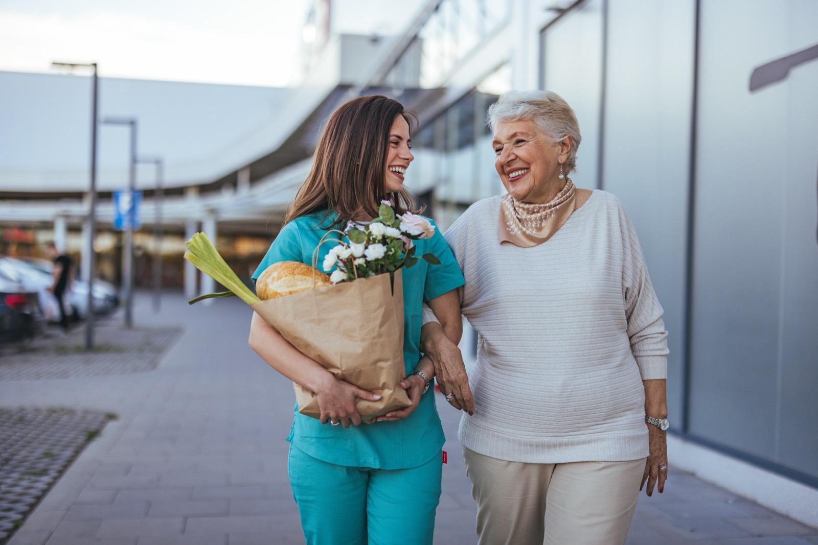 A caregiver in scrubs walks along the street beside an older woman, carrying her groceries and flowers as the two share a laugh.