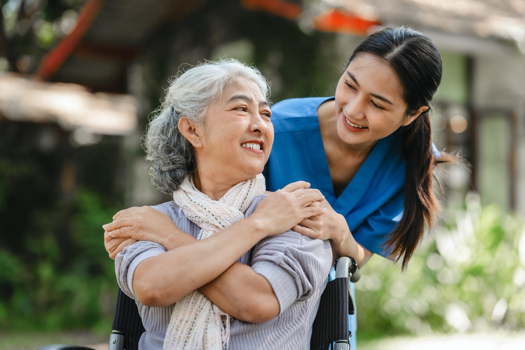 A Gentle Touch caregiver smiles alongside an older woman outdoors, resting a reassuring hand on her shoulder.